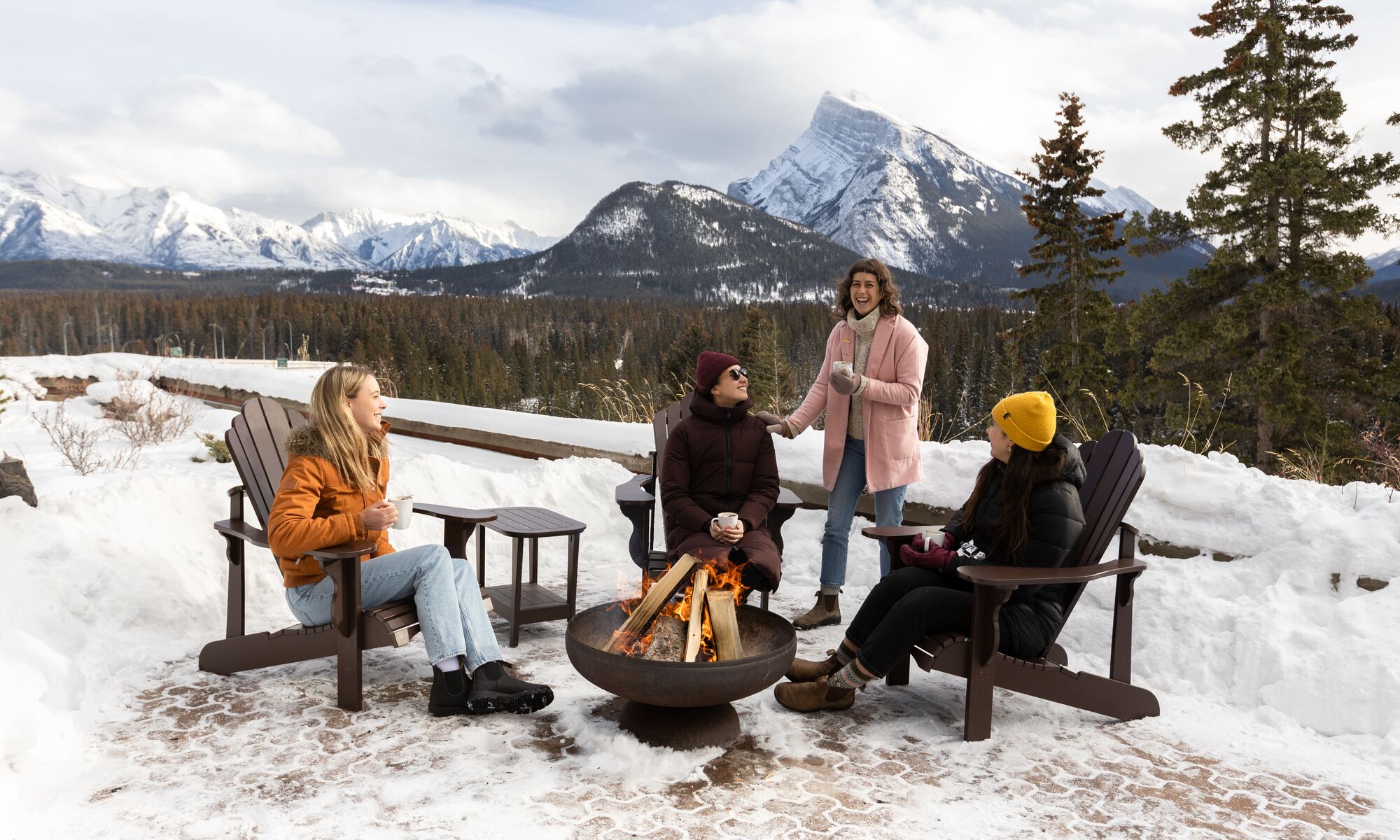 Four friends sit around a campfire in Banff with Rundle Mountain in the background.